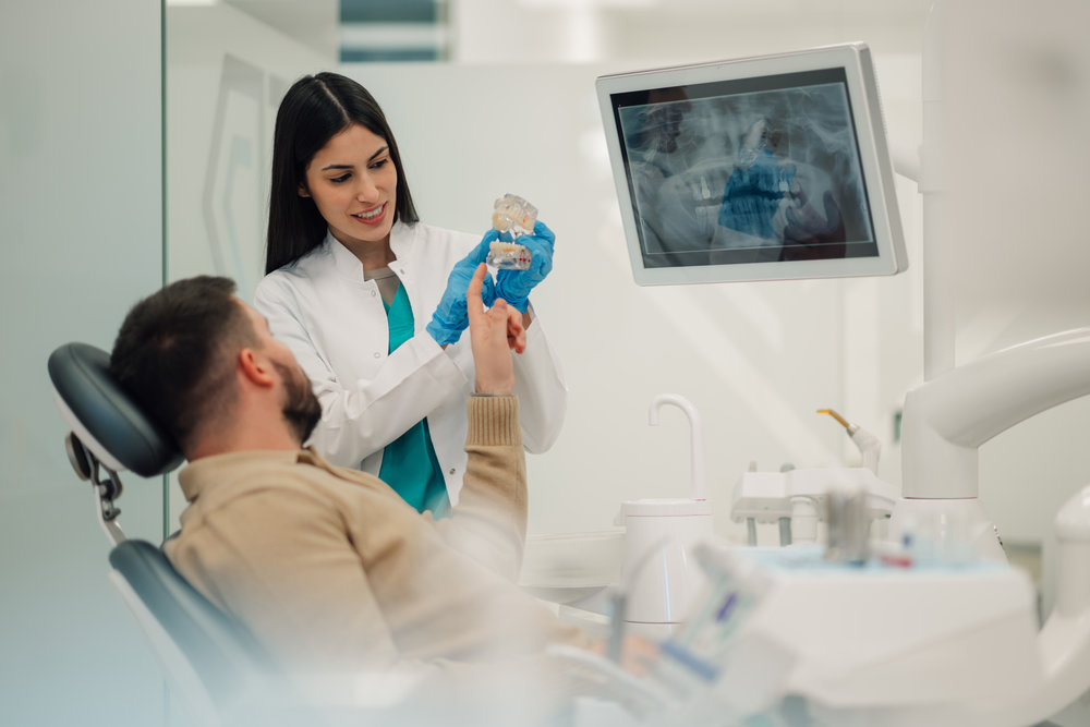 Female dentist is showing a teeth model to a male patient during a consultation in a modern dental clinic. With an x ray image displayed on a screen in the background. Discussing treatment options