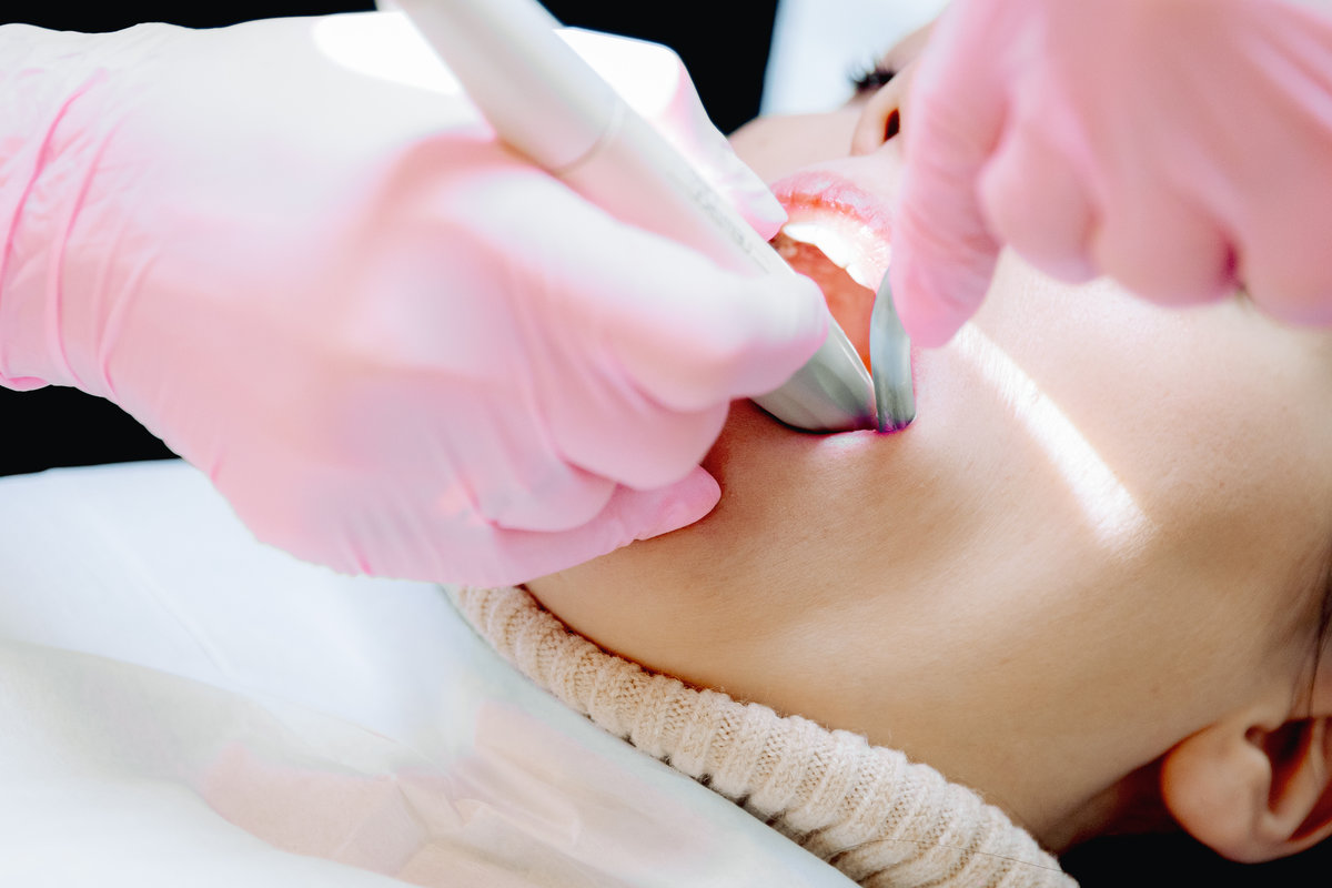 A Caucasian dentist wearing pink gloves cleans tartar from a female patient's left molar with an ultrasonic scaler in her home office during the day, close-up side view. Concept: time to treat teeth, dental procedures, oral health.