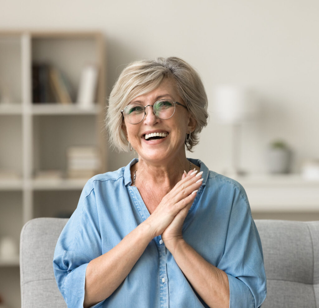 Cheerful pretty older woman in elegant glasses sitting on cozy home couch, smiling with perfect white teeth, laughing with hands at chest gesture, enjoying leisure, comfort, having fun
