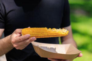 Adult male holding grilled corn on cob in park setting.